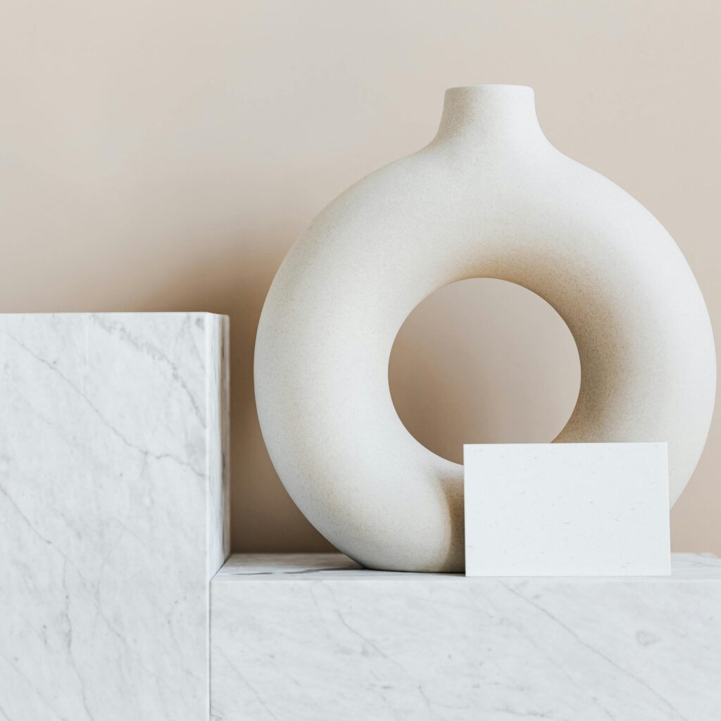 Composition of creative white ceramic vase in ring shape with empty postcard placed on white marble shelf against beige wall as home decoration elements or art objects